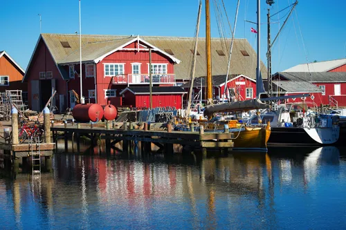 Kleiner Hafen mit roten Holzhäusern und Segelbooten an einem sonnigen Tag.