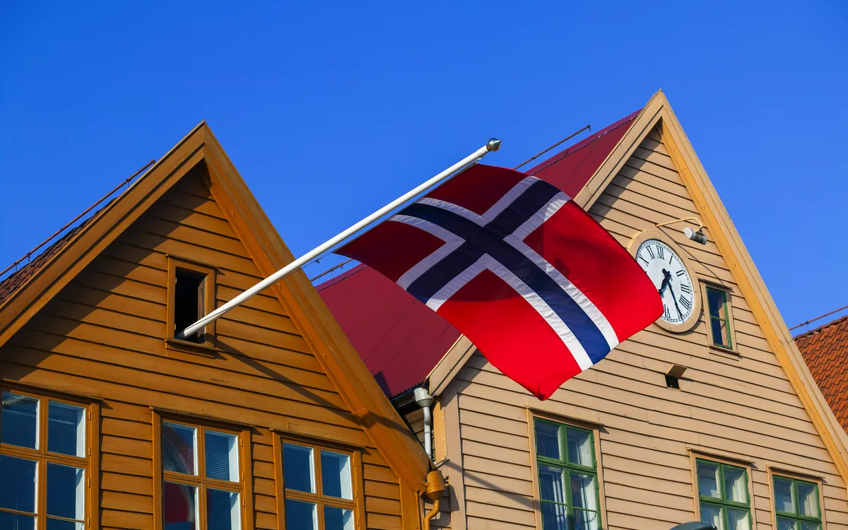 Norwegische Flagge an Holzhäusern mit Uhr und blauem Himmel.