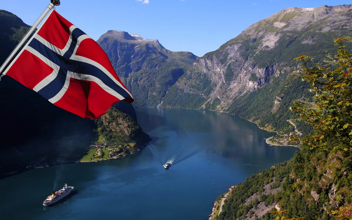 Norwegische Flagge über Fjordlandschaft mit Booten und Bergen im Hintergrund.