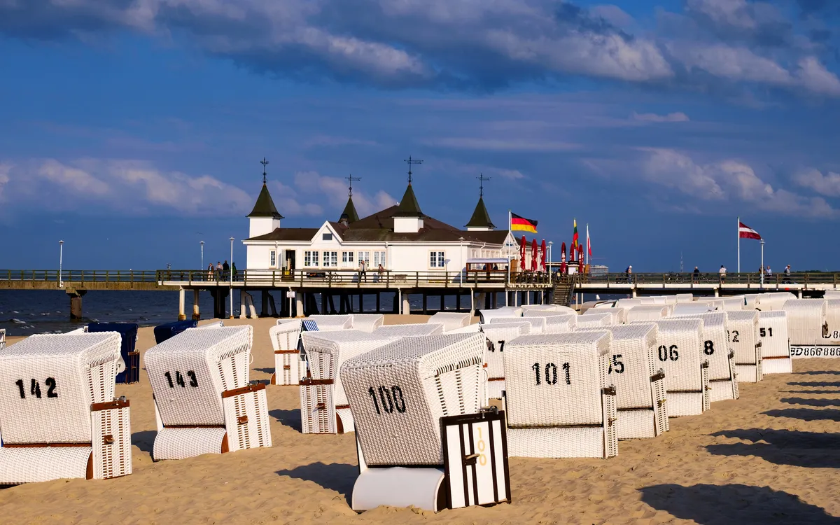 Strandkörbe am Sandstrand vor einer Seebrücke mit historischem Gebäude.