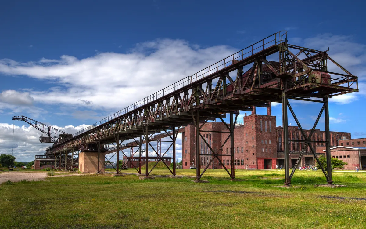Industrieanlage mit Förderband und Backsteingebäude bei blauem Himmel.