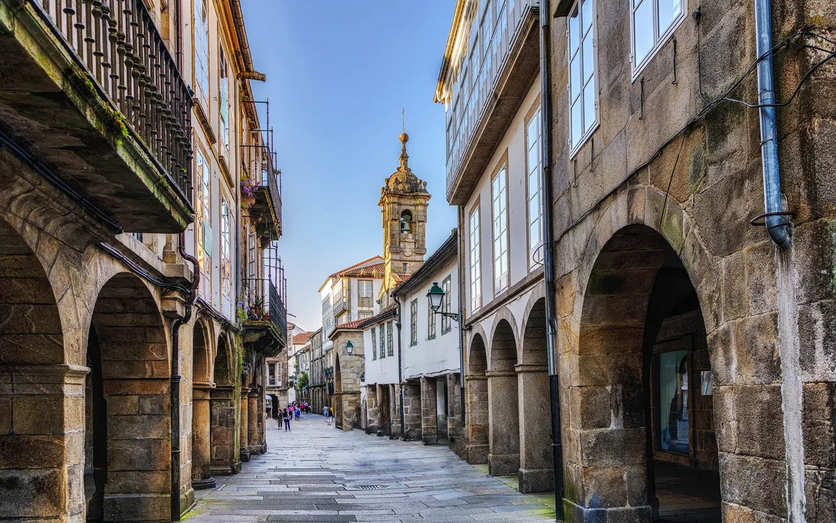 Straße mit historischen Gebäuden und Bogengängen, blauer Himmel im Hintergrund.