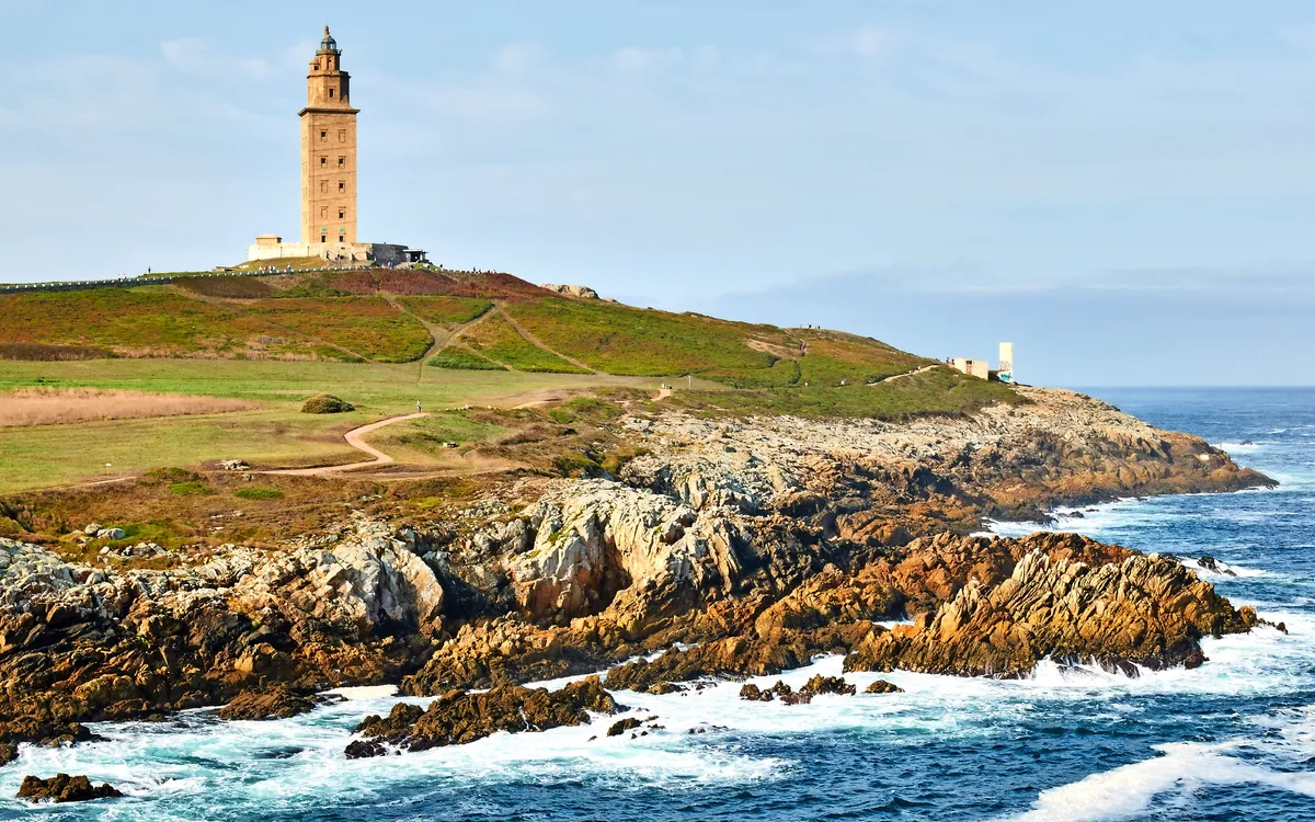 Alter Leuchtturm auf einem Felsen über dem Meer, umgeben von grünen Feldern.