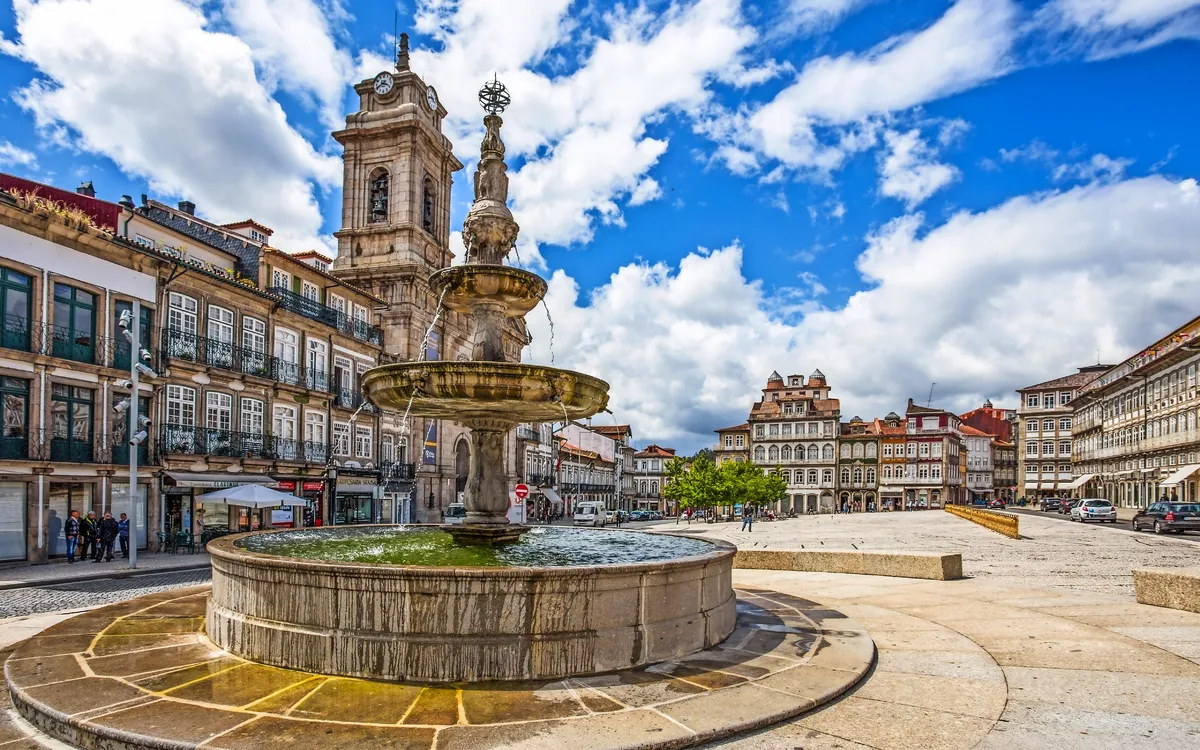 Brunnen auf einem Platz mit historischen Gebäuden und Kirchturm im Hintergrund.