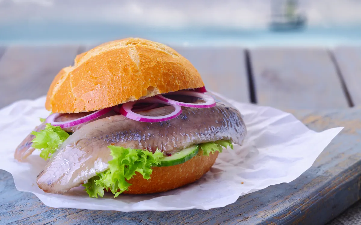Matjesbrötchen mit Hering, Gurken, Salatblatt und Zwiebelringen auf einem Teller vor der Nordsee.