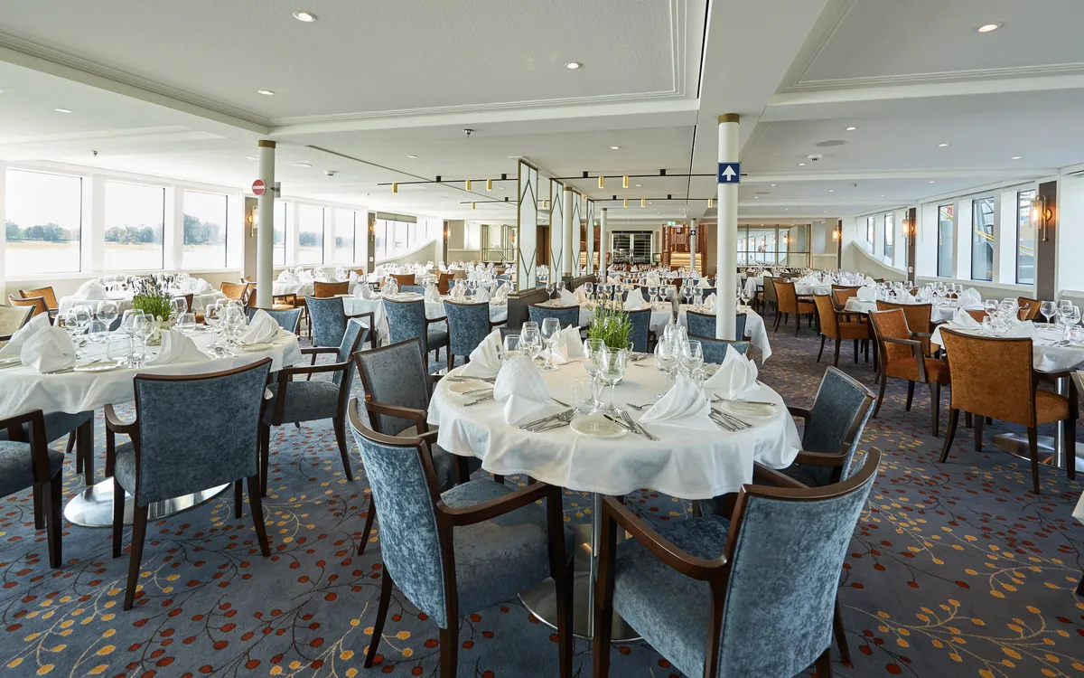 Innenansicht eines elegant gedeckten Kreuzfahrtrestaurants mit runden Tischen und Fenstern.