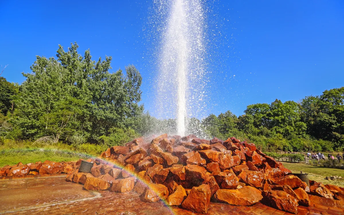 Geysir in Andernach sprüht Wasserfontäne in blauer Himmel, umgeben von Bäumen.