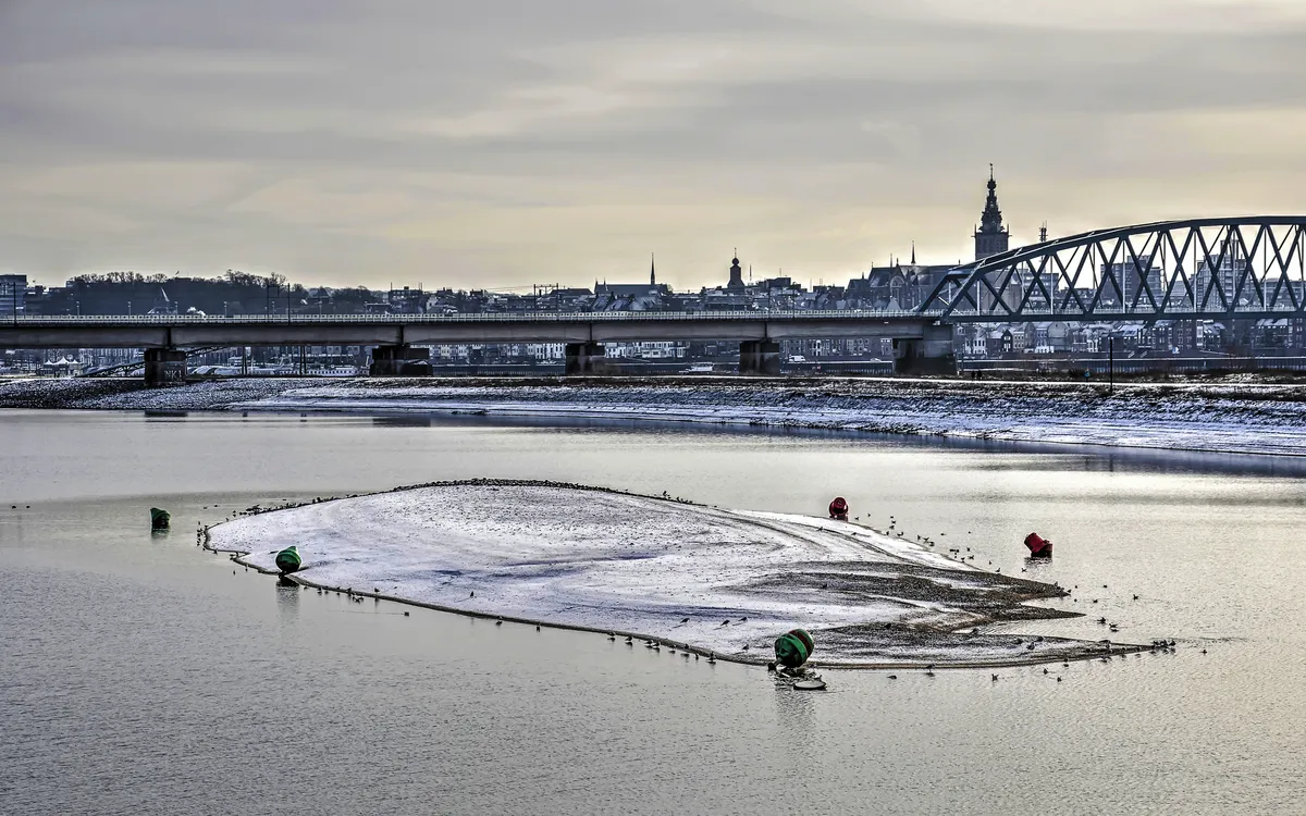 Schneebedeckter Kanal bei Nijmegen, im Hintergrund eine winterliche Stadtlandschaft.