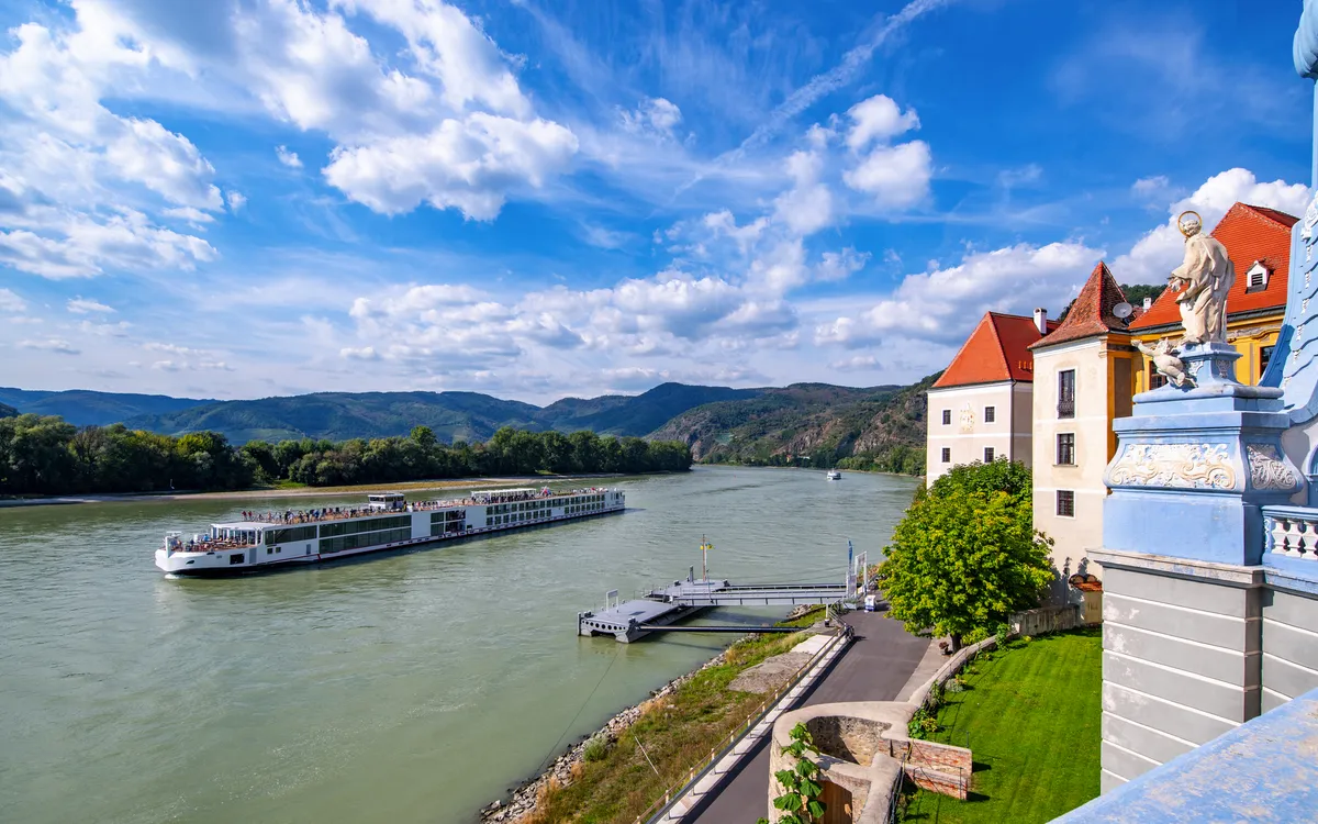 Flusskreuzfahrtschiff auf der Donau vor Schloss und Hügeln unter blauem Himmel.