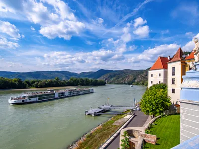 Flusskreuzfahrtschiff auf der Donau vor Schloss und Hügeln unter blauem Himmel.