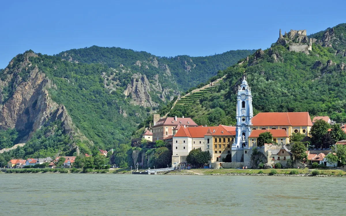 Stadtansicht mit Barockkirche am Fluss vor bewaldeten Hügeln.