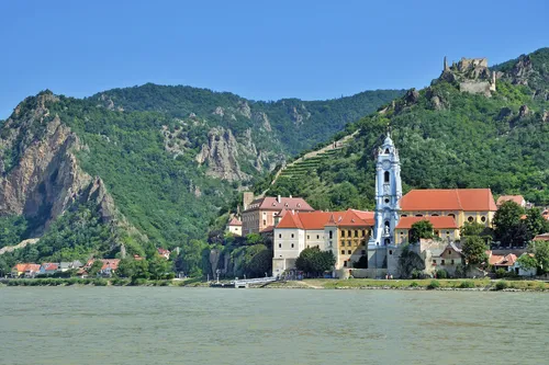 Stadtansicht mit Barockkirche am Fluss vor bewaldeten Hügeln.