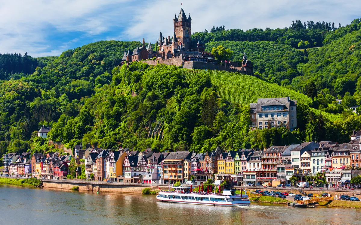 Burg auf Hügel über kleiner Stadt am Flussufer, mit bewaldetem Hintergrund.