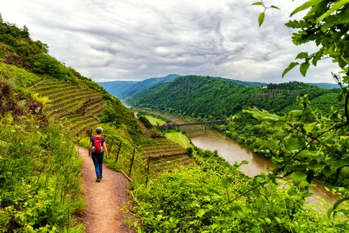 Person wandert auf einem Pfad mit Blick auf Fluss und grüne Hügel.