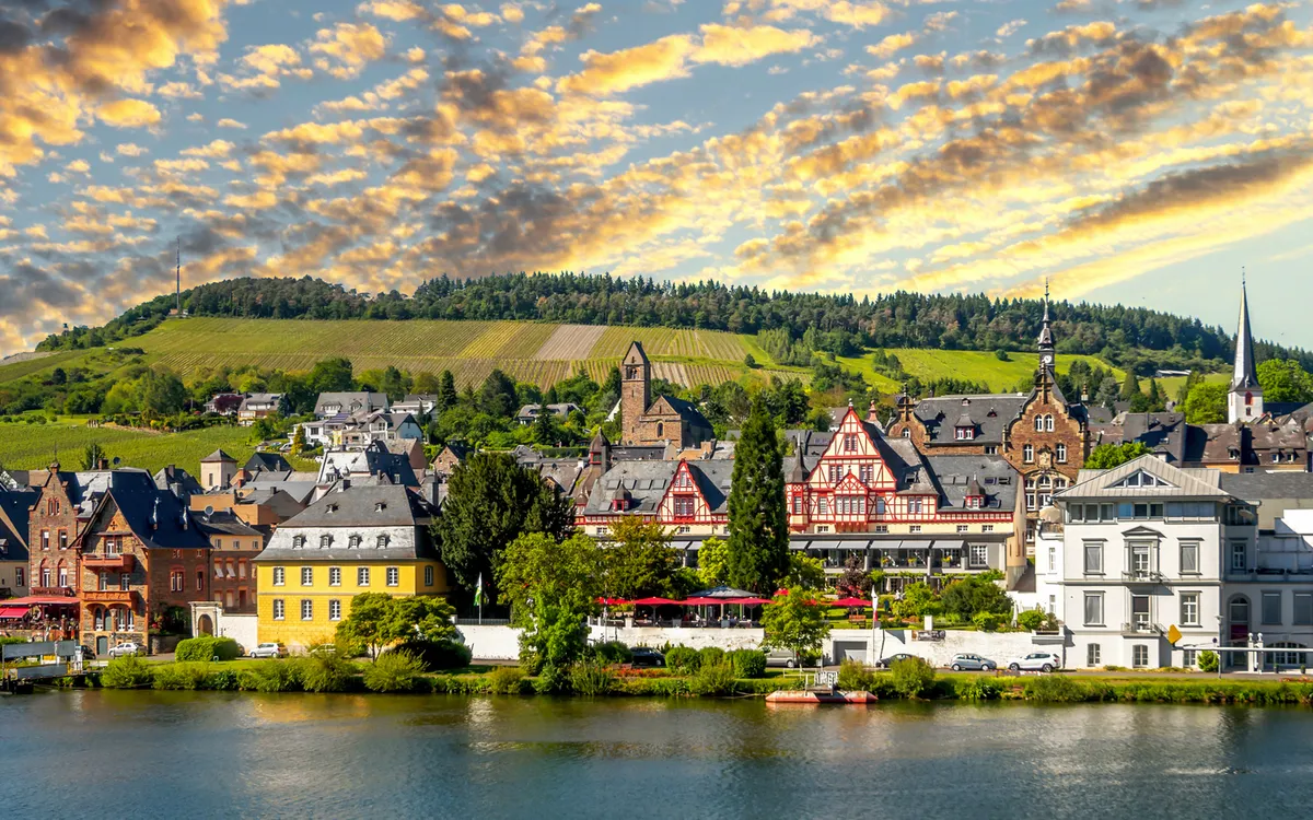 Stadtpanorama mit Fachwerkhäusern an einem Flussufer, im Hintergrund Hügel und bewölkter Himmel.