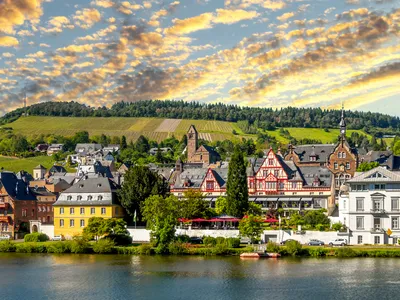 Stadtpanorama mit Fachwerkhäusern an einem Flussufer, im Hintergrund Hügel und bewölkter Himmel.