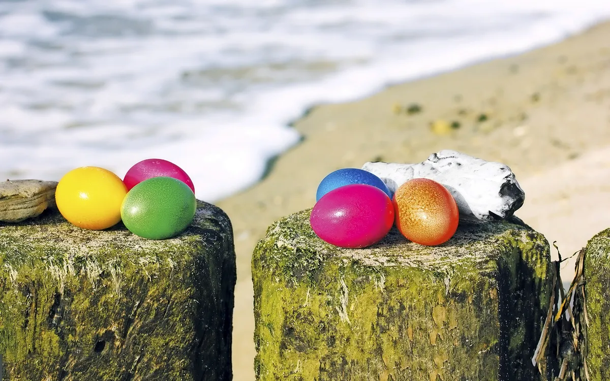 Bunte Ostereier auf Pfählen am Strand mit Meer im Hintergrund.
